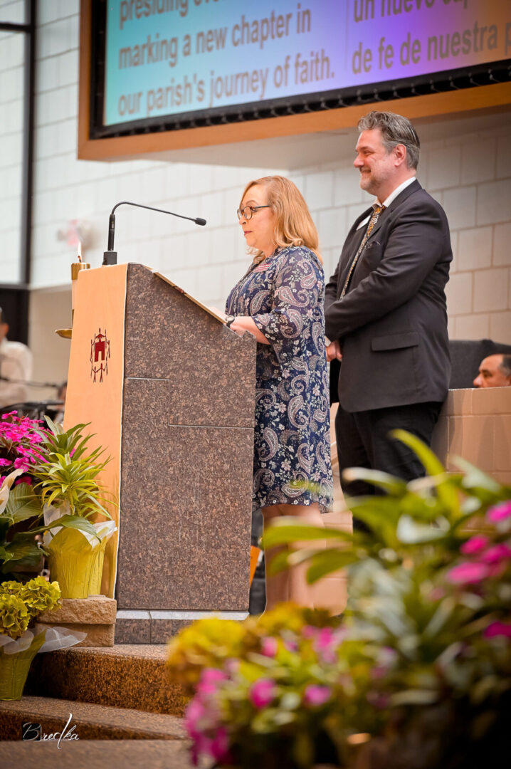 Woman speaking at podium with man standing beside her.